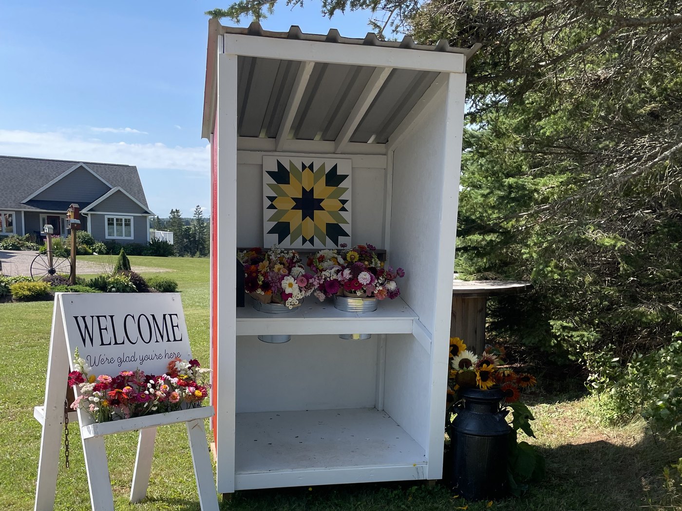 The white roadside flower stand at the end of the lane, its whole roof in frame, bouquets on the shelf and a welcome sign out front